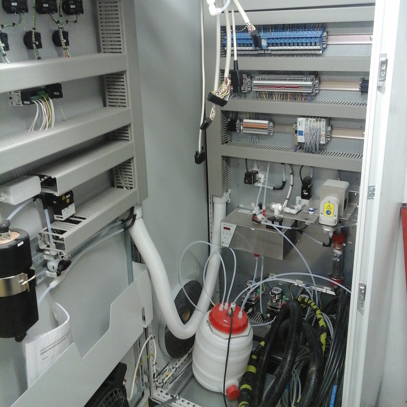 the interior of a control cabinet, likely part of a continuous emission monitoring system (CEMS) in a flue-gas-treatment-plant. The cabinet contains multiple electrical components, including terminal blocks with wiring, relays, and control modules mounted on shelves. There are several tubes and hoses connected to various devices, possibly for gas sampling or calibration. A large white container with a red cap, possibly for calibration gas or liquid, is visible at the bottom, along with neatly bundled cables and a small control panel with gauges and valves. The setup is organized and designed for precise monitoring and control of emissions.
