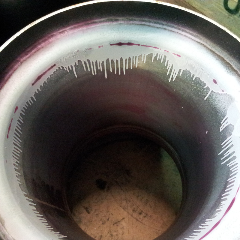 cylindrical metal pipe. The inner surface has a shiny, polished look with some discoloration, showing purple and pink hues near the top edge. There are also white, drip-like marks running down from the edge, possibly indicating some residue or contamination. The bottom of the pipe appears darker and less reflective.