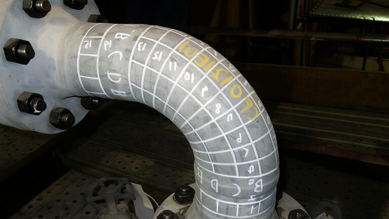 metal pipe elbow with a grid pattern marked on its surface. The grid appears to be part of a non-destructive testing procedure, most likely ultrasonic thickness measurements (UT), used to assess the wall thickness and structural integrity of the pipe. Within the grid, there are various numbers and letters, which likely represent measurement values or specific inspection codes. A prominent yellow marking "F0121E01" is visible, which serves as an identifier for the pipe or inspection area. The pipe is bolted to a flange, indicating that it is part of a pressurized piping system, probably used in an industrial environment such as a refinery, chemical plant, or power station.