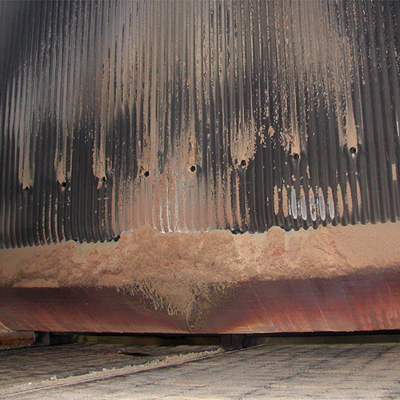 heat exchanger or similar equipment. It features a series of vertical metal tubes or fins, covered with rust, soot, and a significant amount of accumulated dust or ash. The lower part of the image reveals a buildup of debris or sediment at the base, suggesting prolonged use or lack of maintenance. The metal surfaces show signs of wear and corrosion, with several small holes visible along the tubes. The overall appearance indicates an aged industrial component, possibly requiring cleaning or repair.