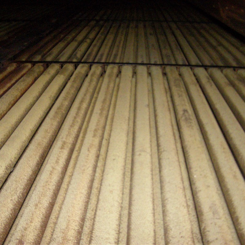interior of a heat exchanger, likely part of a Heat Recovery Steam Generator (HRSG). Visible are parallel heat exchanger tubes, possibly evaporator or superheater tubes. The tubes are covered with deposits and rust, indicating prolonged use in harsh thermal and chemical conditions. The lighting is dim, suggesting the photo was taken in an enclosed space, such as an HRSG gas duct. The structure is made of metal with visible cross supports.