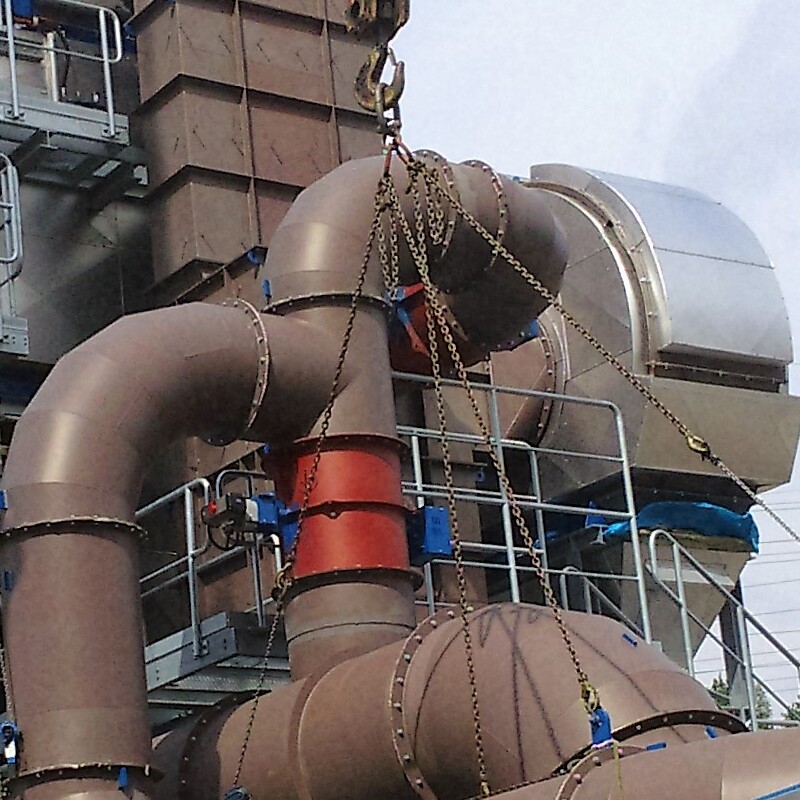 section of a large industrial installation, likely during assembly. At the center is a system of pipes and components, including large, rounded metal elements with a red and blue valve. The pipes and parts are supported by chains and a hook, suggesting they are being lifted or positioned. In the background, there are metal structures, platforms, and railings, with a glimpse of the sky, indicating the installation is outdoors or in an open hall. The setup appears to be part of a larger system, such as in a power plant or industrial facility.