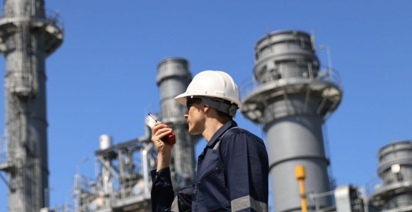 industrial worker wearing a white safety helmet and a blue uniform with reflective strips, standing at a large industrial facility. The worker is holding a walkie-talkie and appears to be communicating. In the background, there are several tall cylindrical structures, likely part of a refinery or power plant, with metal staircases and railings. The sky is clear and blue, indicating a sunny day.