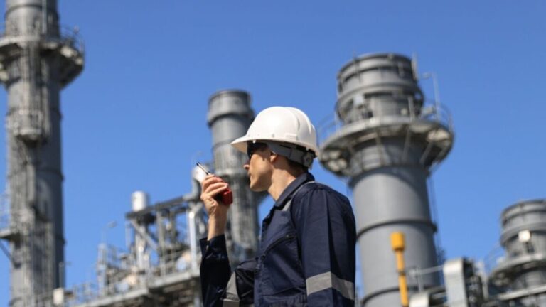 industrial worker wearing a white safety helmet and a blue uniform with reflective strips, standing at a large industrial facility. The worker is holding a walkie-talkie and appears to be communicating. In the background, there are several tall cylindrical structures, likely part of a refinery or power plant, with metal staircases and railings. The sky is clear and blue, indicating a sunny day.