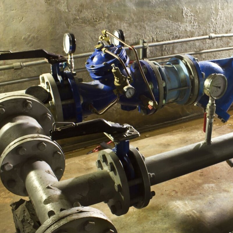 section of an industrial piping system, likely in a utility or mechanical room. It features large metal pipes connected with flanges and bolts, along with blue valves and fittings. There are two pressure gauges mounted on the system, one on a blue valve and another on a pipe, for monitoring pressure levels. The setup includes various control valves with handles, indicating manual operation. The background shows a rough concrete wall, and the floor appears to be concrete as well, suggesting a rugged, functional environment typical of industrial facilities.