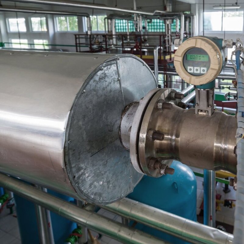 the interior of an industrial facility, likely a production plant or laboratory. At the center is a large metal setup—a cylindrical tank or pipe, connected to other components with flanges and bolts. A digital pressure or flow meter is mounted on the setup, displaying values on its screen. In the background, there are pipes, valves, blue tanks, and other industrial equipment. The room has large windows letting in natural light, and white walls with green accents. The space appears to be a well-organized workplace related to technical processes.