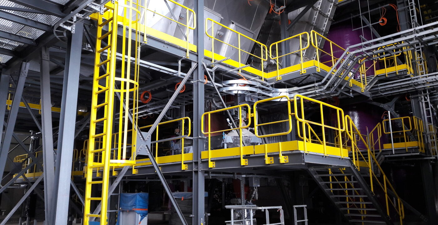 a section of a flue-gas-treatment-plant. It features a multi-level industrial structure with metal platforms, yellow safety railings, and ladders for access. Large metallic ducts and equipment, likely for filtering or treating flue gases, are visible. The setup includes pipes, valves, and various mechanical components, all within a robust steel framework, indicating a focus on safety and functionality in an industrial environment.
