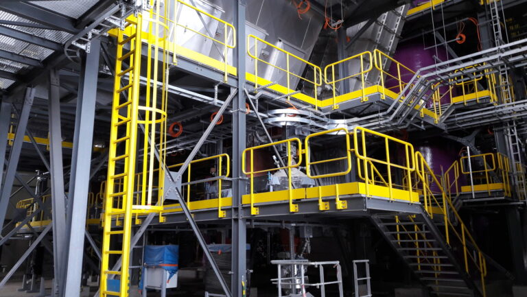 a section of a flue-gas-treatment-plant. It features a multi-level industrial structure with metal platforms, yellow safety railings, and ladders for access. Large metallic ducts and equipment, likely for filtering or treating flue gases, are visible. The setup includes pipes, valves, and various mechanical components, all within a robust steel framework, indicating a focus on safety and functionality in an industrial environment.