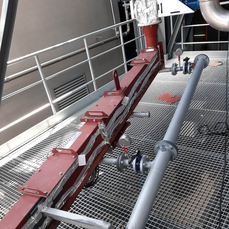 "A fluidized transport duct for residue dust, painted red-brown with primer paint. Trace heating is visible on the walls. A fresh fluidizing air pipe is attached to the side. The duct features multiple inspection windows. The supply pipes are equipped with visible manual shut-off valves and pressure gauges. The setting is industrial, with a galvanized safety railing in the background and grating walkway panels on the floor