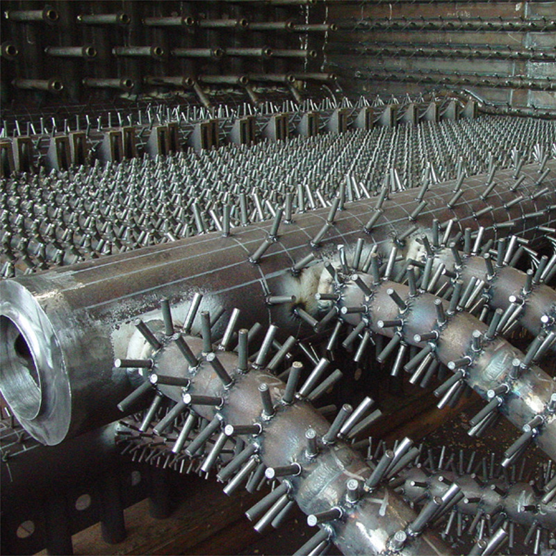 interior of an industrial heat exchanger or similar equipment. It features multiple rows of metal tubes, some of which are covered with numerous small, evenly spaced metal fins or studs welded onto them. These fins are likely designed to increase the surface area for heat transfer. The tubes and surrounding structure appear to be made of steel or another industrial metal, with visible rust and wear, indicating prolonged use. The background includes additional layers of tubes and a sturdy framework, suggesting a complex and robust design typical of heavy industrial machinery.
