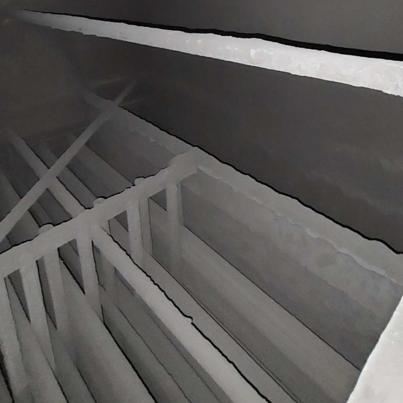 The gas chamber filter’s interior is dusty, with fine ash in the air. Rows of fabric filter bags, coated with particles, hang over metal cages, capturing dust from flue gas. The walls are corrosion-resistant, and walkways allow maintenance. The air is heavy.