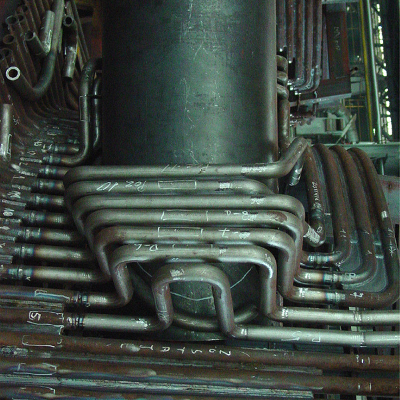 industrial heat exchanger or similar equipment. It features multiple rows of metal tubes, some arranged in U-shaped bends, densely packed together. The tubes are made of steel or a similar material and show signs of rust and wear, with visible weld marks and handwritten labels or markings on them. The central part of the image includes a large vertical cylindrical component, possibly a central support or another section of the exchanger. The overall appearance suggests a complex and robust design, typical of heavy industrial machinery used for heat transfer processes.