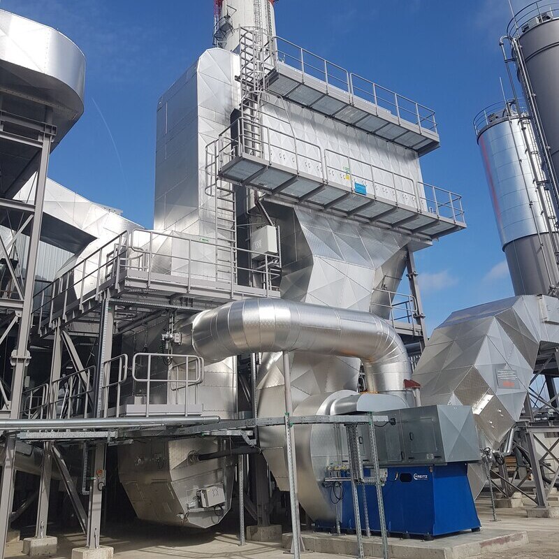 The image shows a flue gas treatment installation, part of an industrial facility used for cleaning exhaust gases from a combustion process. The entire structure is covered in metallic, insulated cladding to handle high-temperature gases and ensure thermal efficiency. At the center of the image is a large recirculation duct connected to a high-capacity industrial fan, which is enclosed in a blue housing. This fan forces the flue gases through the system. Above the duct, there is a vertical chamber with inspection platforms and access ladders, indicating this is the main filtration or separation unit where particles and pollutants are removed from the gas stream. A tall chimney at the top of the structure is used for releasing the treated gases into the atmosphere. The entire setup is engineered for environmental compliance and is designed to operate continuously under demanding industrial conditions.