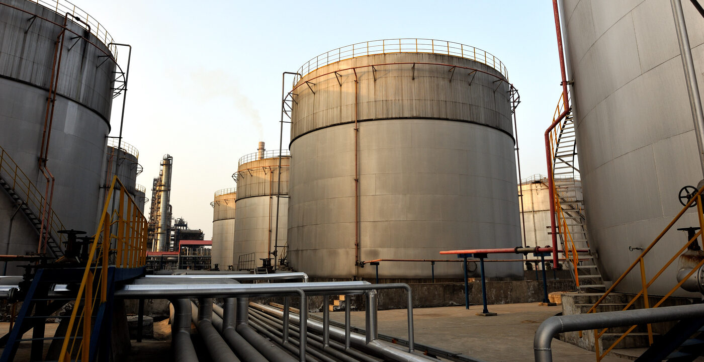 an industrial facility with several large cylindrical storage tanks made of metal, likely used for storing liquids such as oil, chemicals, or gas. The tanks are equipped with staircases, railings, and pipes, indicating a complex infrastructure. Some pipes are elevated and run horizontally across the ground, while others connect to the tanks. The facility appears to be operational, with visible smoke or vapor rising from some areas, suggesting active processing or storage. The scene is set during daylight with a clear sky in the background.