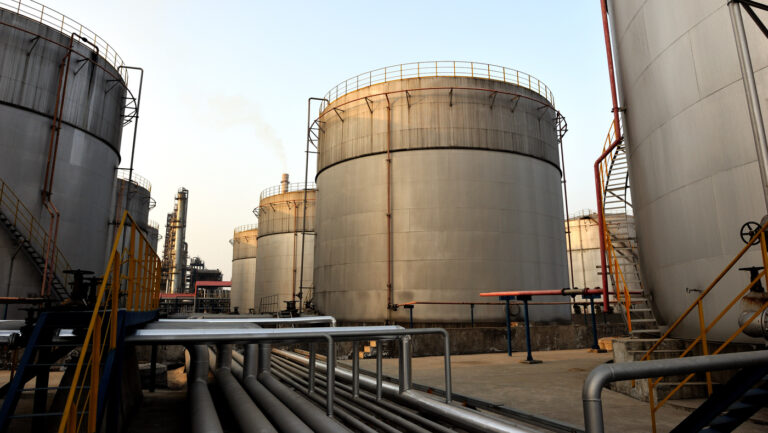 an industrial facility with several large cylindrical storage tanks made of metal, likely used for storing liquids such as oil, chemicals, or gas. The tanks are equipped with staircases, railings, and pipes, indicating a complex infrastructure. Some pipes are elevated and run horizontally across the ground, while others connect to the tanks. The facility appears to be operational, with visible smoke or vapor rising from some areas, suggesting active processing or storage. The scene is set during daylight with a clear sky in the background.