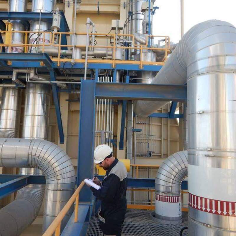 Power plant. There are large, insulated metal pipes and ducts, some labeled with "Steam" and directional arrows. A worker wearing a hard hat and safety gear is standing on a grated platform, holding a clipboard and taking notes. The structure includes blue metal frameworks with yellow railings, and various equipment and piping systems are visible in the background, indicating a complex industrial process involving steam or fluid transport.