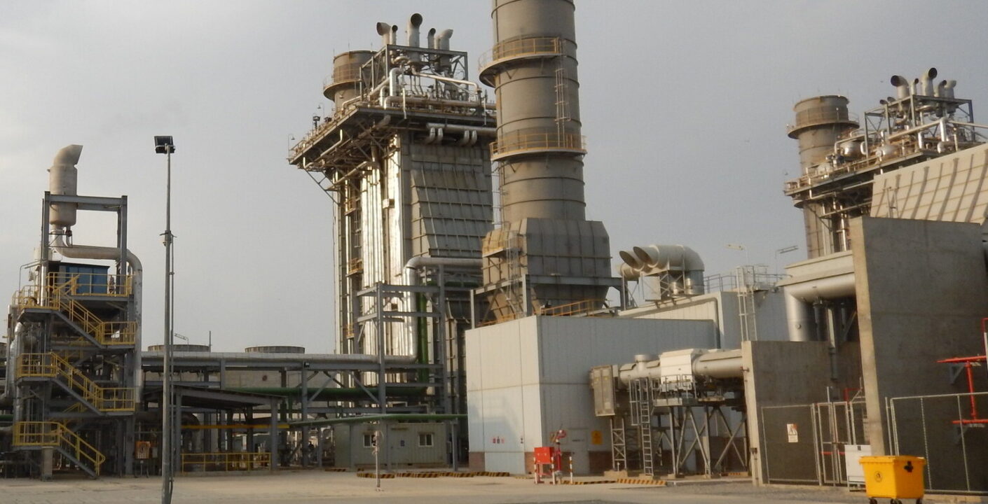 industrial facility, likely a power plant or refinery. It features several large cylindrical towers and chimneys, with extensive piping and metal frameworks. The structures include staircases, platforms, and safety railings, indicating a complex operational environment. There are various industrial components such as ducts, valves, and control units. A yellow waste bin and a fenced area are visible in the foreground, and the sky is overcast with some clouds. The overall setting appears to be an outdoor industrial site with heavy machinery and infrastructure.