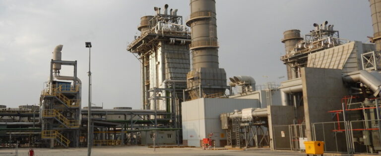 industrial facility, likely a power plant or refinery. It features several large cylindrical towers and chimneys, with extensive piping and metal frameworks. The structures include staircases, platforms, and safety railings, indicating a complex operational environment. There are various industrial components such as ducts, valves, and control units. A yellow waste bin and a fenced area are visible in the foreground, and the sky is overcast with some clouds. The overall setting appears to be an outdoor industrial site with heavy machinery and infrastructure.