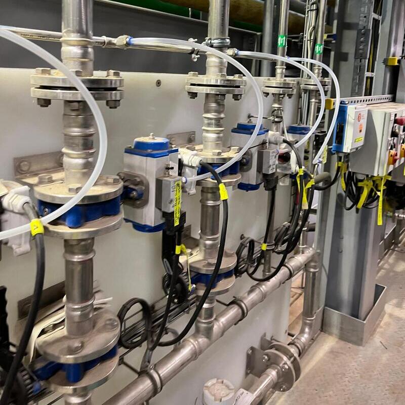 an industrial installation, likely in a factory or processing plant. Visible are metal pipes, valves, and measuring devices such as pressure gauges and sensors. Blue devices with displays, possibly pressure or flow transmitters, are mounted on the pipes. There are also transparent tubes, cables, and a control panel with buttons on the right side. The setup appears to be part of a system for handling fluids or gases.