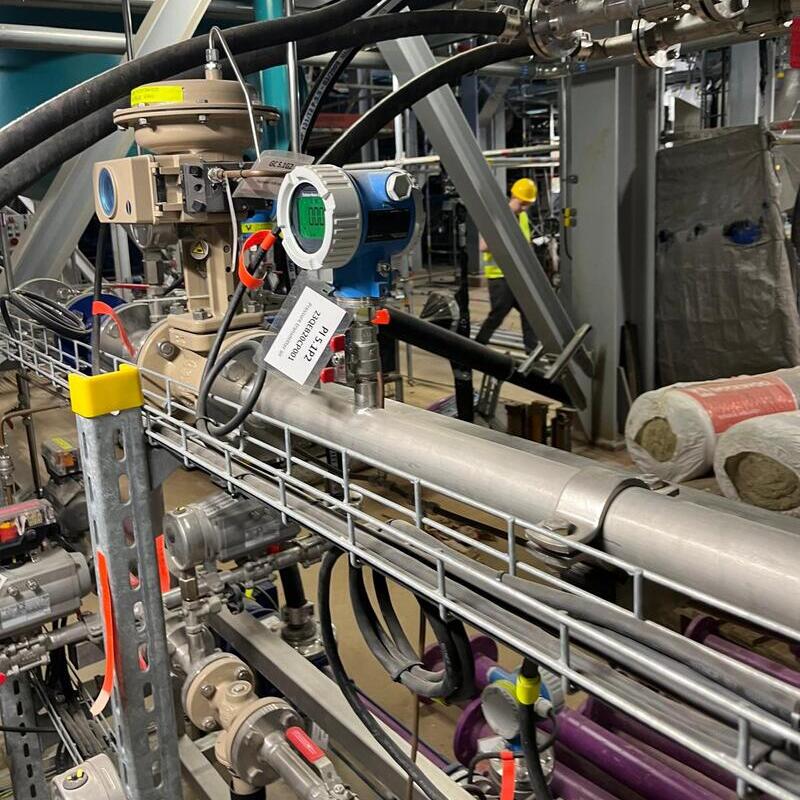 an industrial setting, likely a factory or processing plant. There are large metal pipes, valves, and pressure gauges visible, along with various cables and hoses. A digital pressure transmitter with a green display is mounted on the pipe, showing a reading of "0.00." A tag attached to the equipment reads "PID 1000A5 T4F." In the background, there are workers wearing safety helmets and vests, and some insulation materials are stacked on the right side. The environment appears to be focused on handling fluids or gases, typical of a chemical or manufacturing facility.