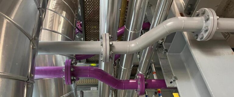 a section of an industrial setup, likely in a chemical or processing plant. There are pipes of various diameters, some painted purple and others gray. The pipes are connected with flanged joints secured by bolts, indicating a system that requires tight sealing. In the background, there are larger metal components, possibly tanks or ventilation ducts, along with supporting structures like metal beams. The setup appears to be a well-organized technical infrastructure.