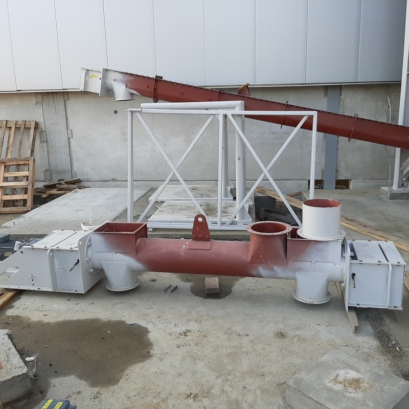 "A screw conveyor for residue dust with a circular cross-section, featuring two outlets and one inlet, a console for the electric drive, and bearing supports. It is painted with red-brown primer paint. In the background, another screw conveyor with a V-shaped design is visible, mounted on a supporting structure, also painted in red-brown