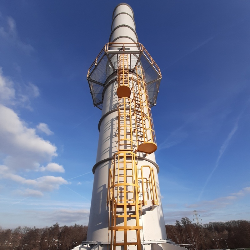 cylindrical industrial stack made of a light-colored steel. On its sides, there are yellow ladders with safety railings leading to platforms at different levels. The background features a blue sky with a few clouds, and at the base of the chimney, there are leafless trees, indicating winter or early spring. In the distance, power lines and a transmission tower are visible.