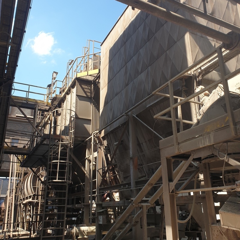 a zinc flue-gas-treatment-plant. It features a multi-level structure with metal platforms, railings, and ladders for access to various sections. At the center, there are large tanks and ducts, likely used for filtering or treating flue gases, such as zinc dust, SO₂, or other pollutants. In the background, a large tank wall with a geometric pattern is visible, and the entire setup is well-illuminated by natural light, indicating an outdoor or semi-outdoor industrial environment.